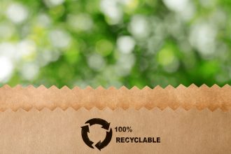 Blurred view of green tree outdoors. Bokeh effect Paper bag with recycling symbol on blurred green background. Eco friendly package